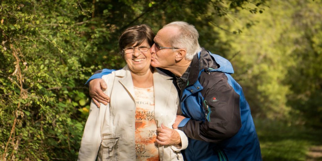 An elderly man kissing a smiling elderly woman on the cheek.